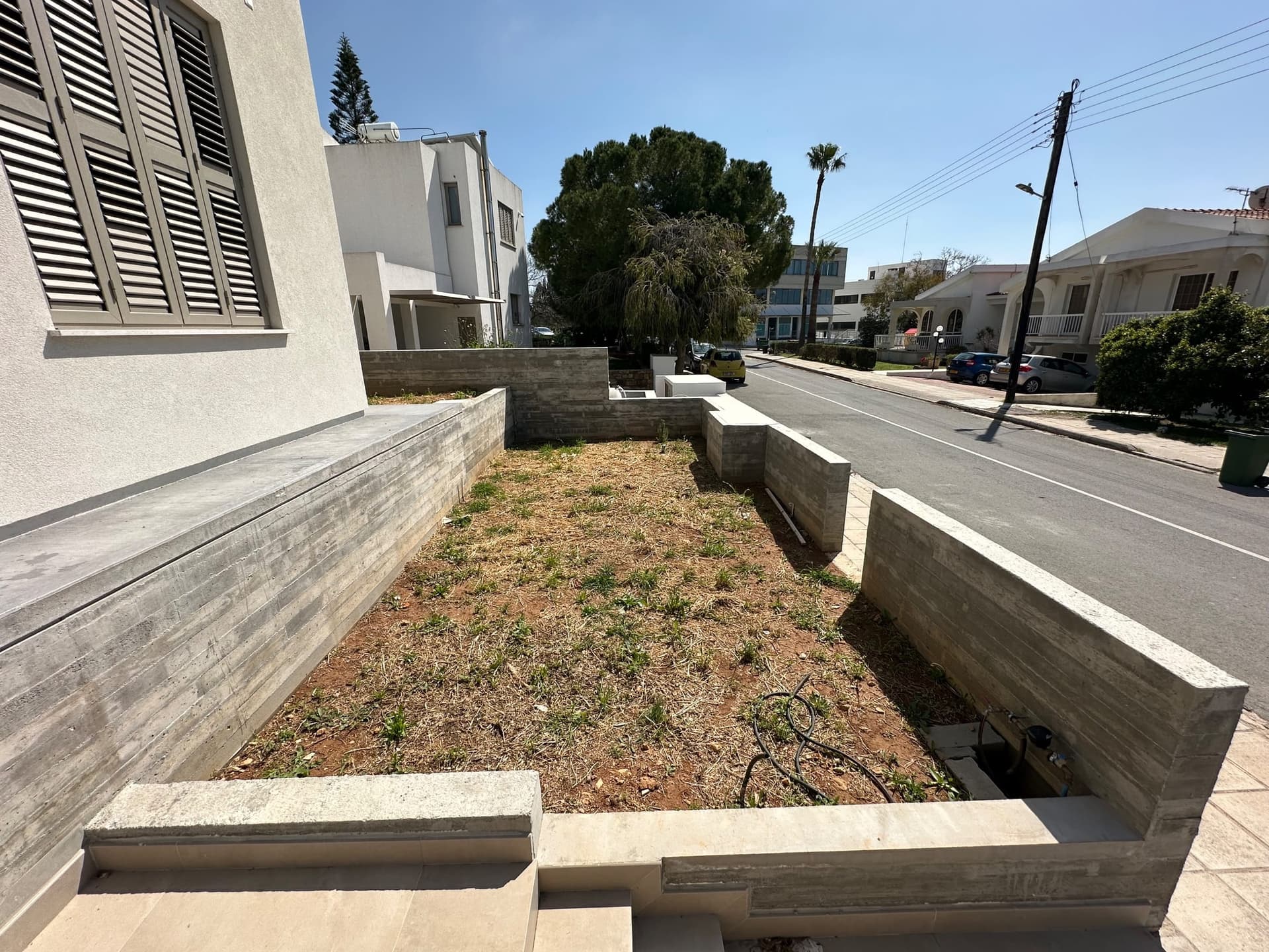 Narrow front garden bed with concrete retaining walls and sparse plantings next to white modern house and street.