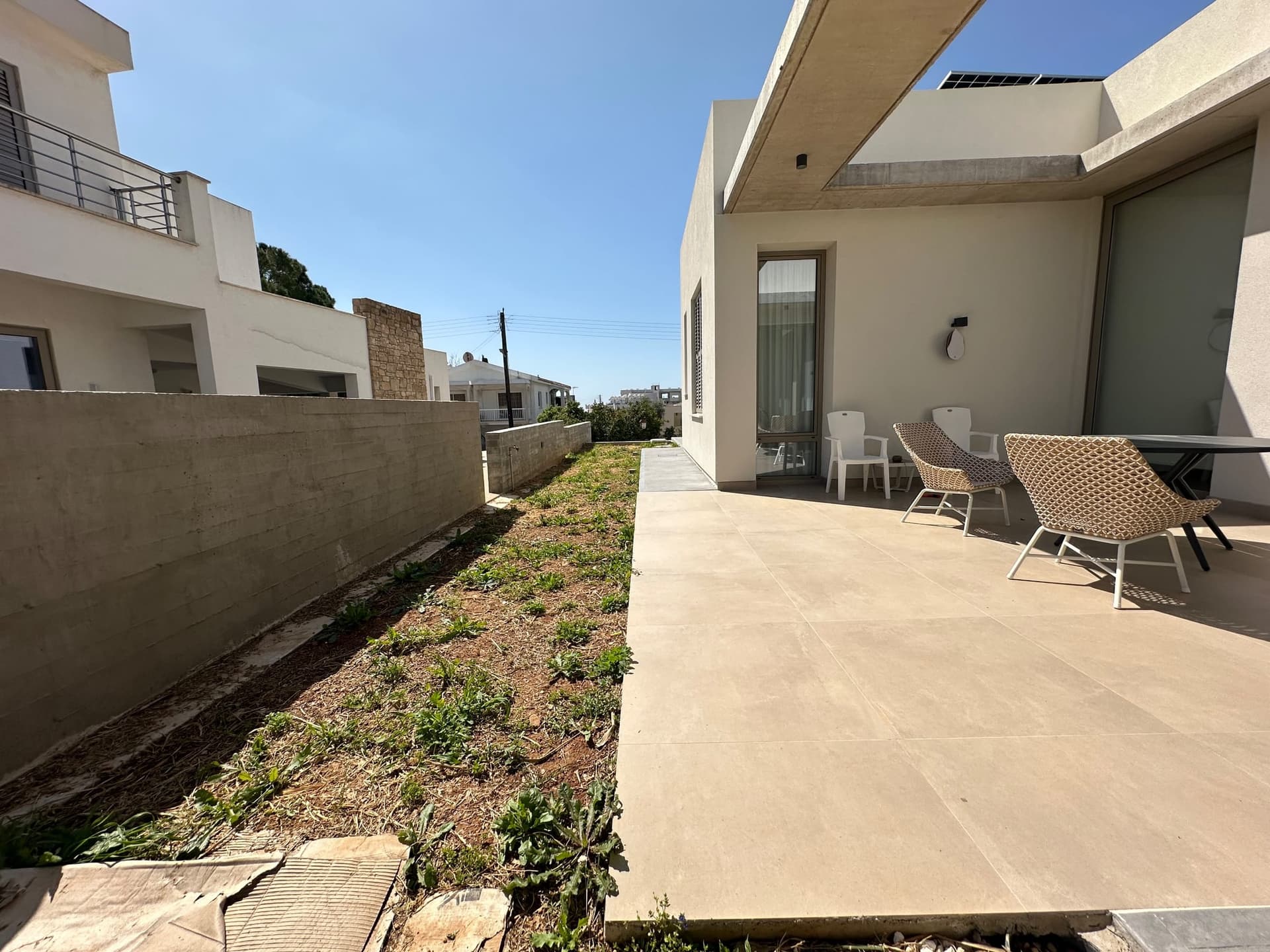 Modern residential garden with concrete patio, contemporary white chairs, concrete boundary wall, and newly planted groundcover alongside contemporary home architecture.