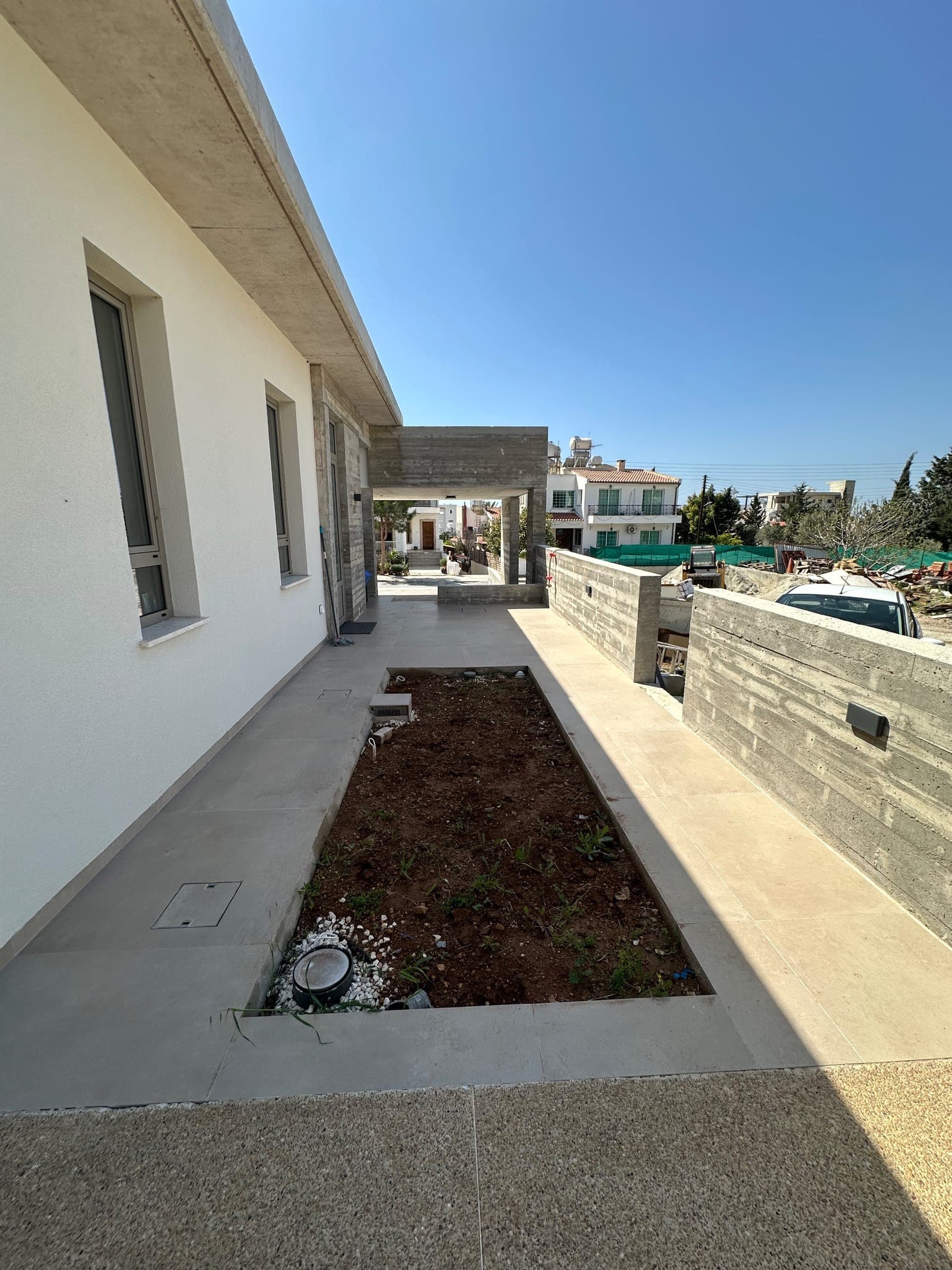 Narrow raised garden bed with dark soil along a modern paved terrace beside a white house, with stone retaining walls and Mediterranean landscape beyond.