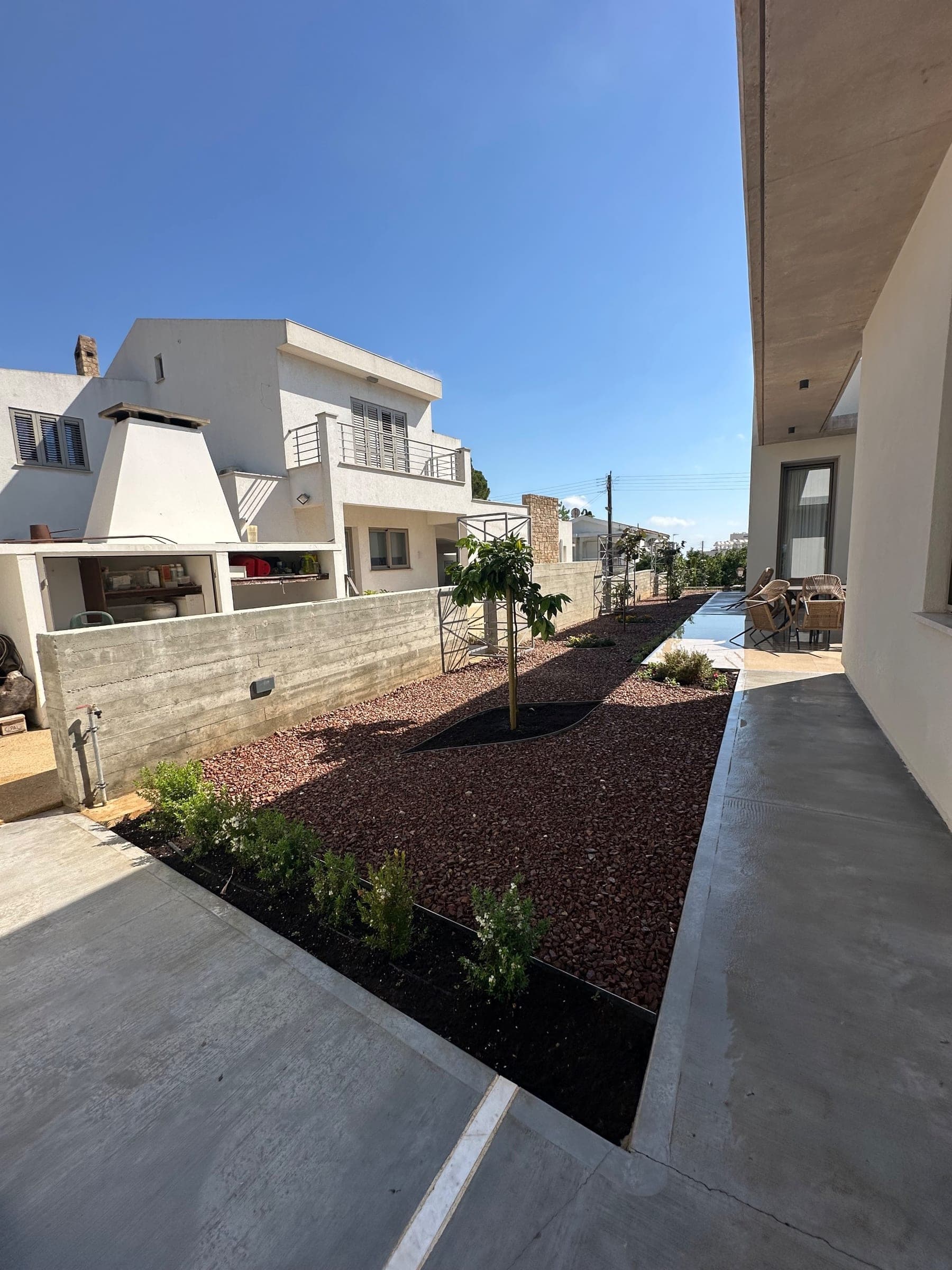 Modern Mediterranean garden with reddish-brown gravel beds, young tree plantings, low black steel edging, concrete paving, and contemporary residential architecture.