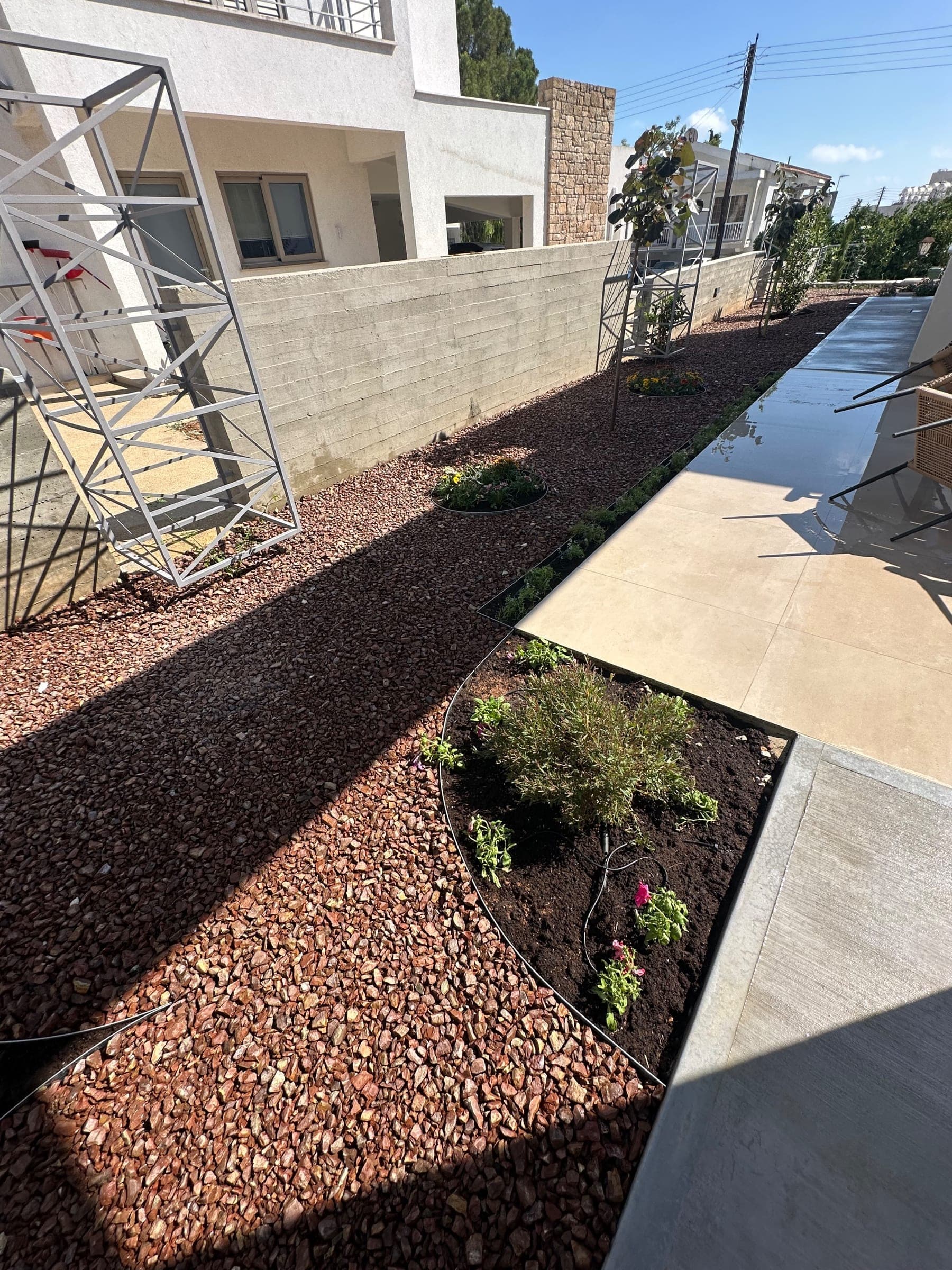 Modern Mediterranean garden with reddish-brown gravel beds, young shrubs, concrete paving, rendered walls, and climbing structures against contemporary architecture.