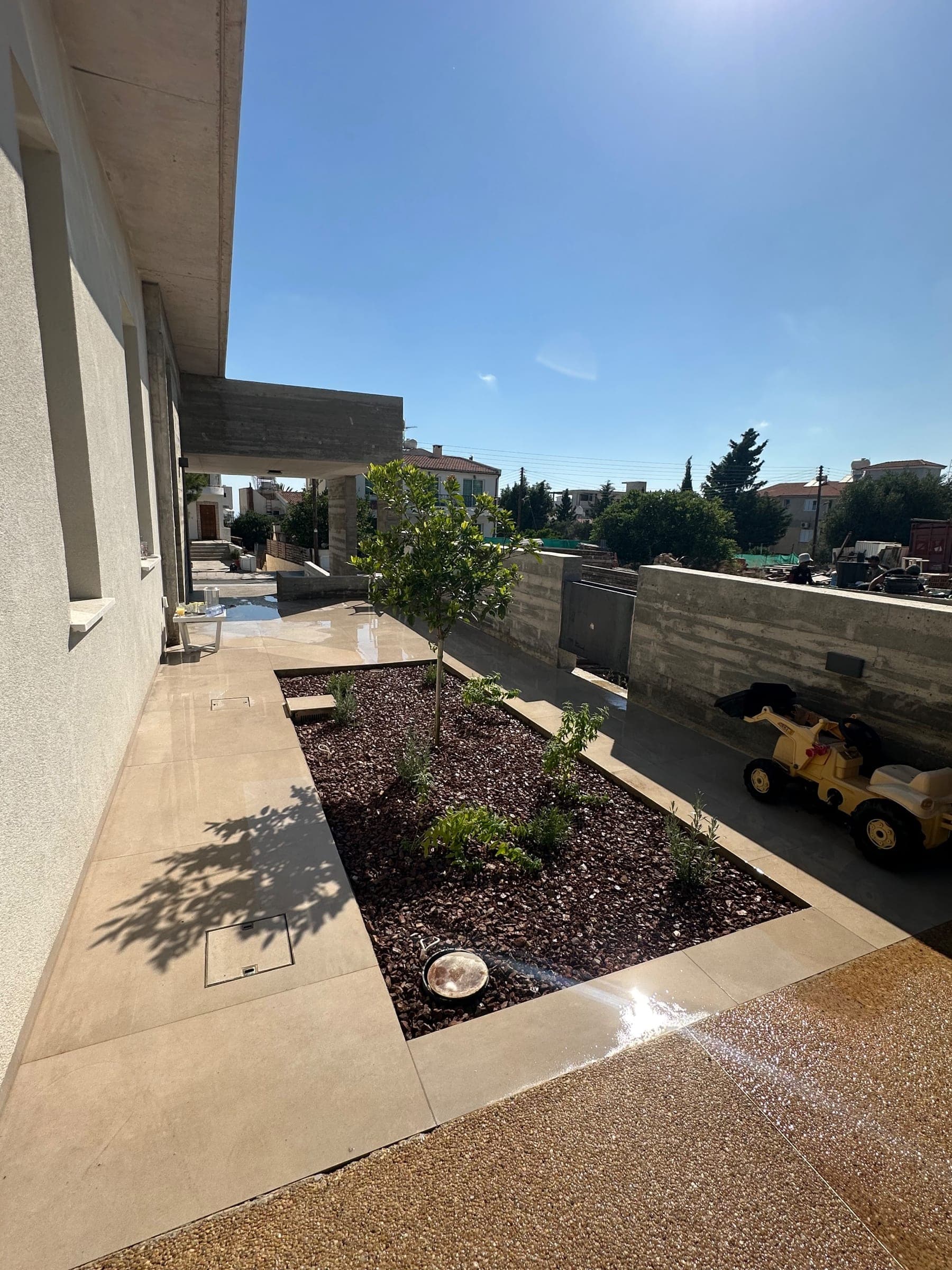 Rooftop terrace with paved flooring, dark gravel planting bed with young tree, concrete walls, and Mediterranean townscape views under clear sky.
