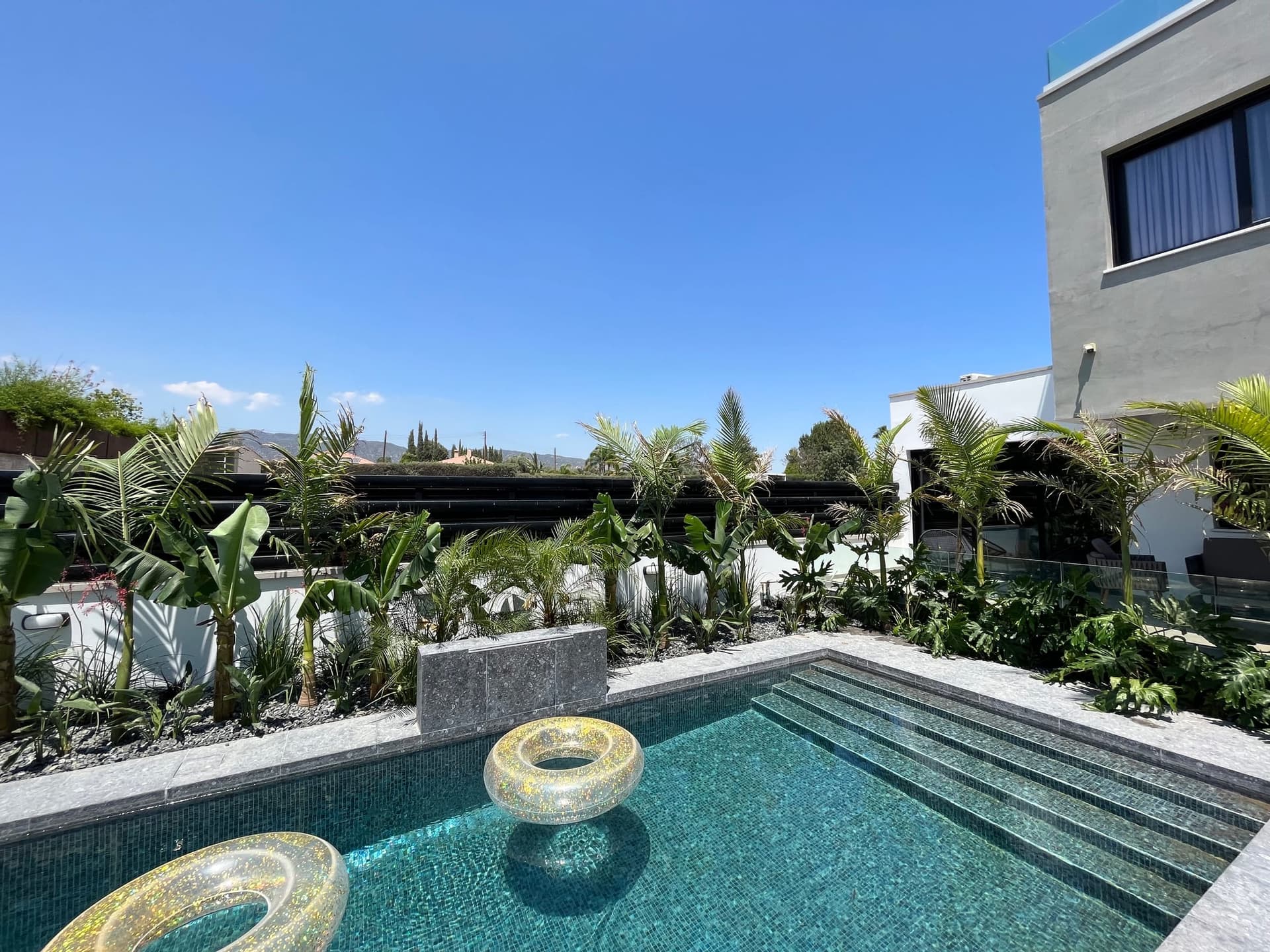 Modern pool courtyard with palm trees, tropical plants, and gray stone hardscaping bordering a tiled swimming pool, backed by pergola and contemporary home.