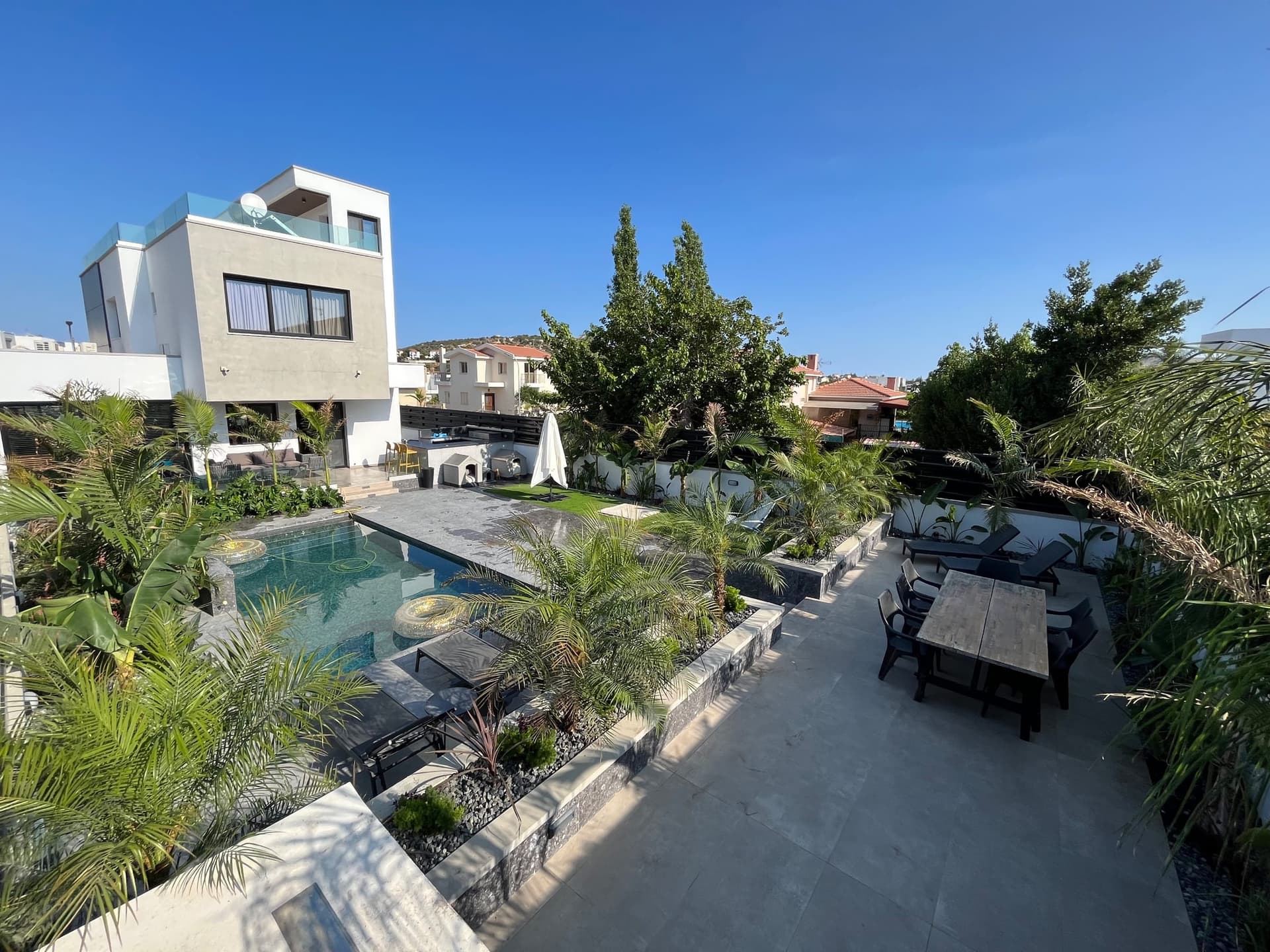 Modern villa garden with swimming pool, paved terrace, dining area with black loungers, and tropical palm planting.