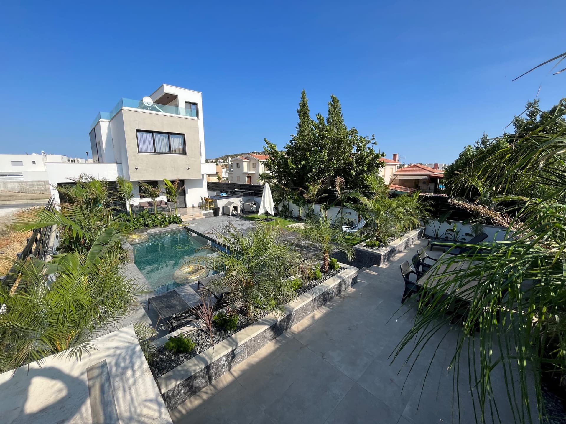 Modern courtyard garden with swimming pool, paved terracing, palm trees, and black lounge seating surrounded by Mediterranean landscaping.